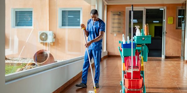 A man in blue scrubs mopping a tiled floor with cleaning supplies nearby.