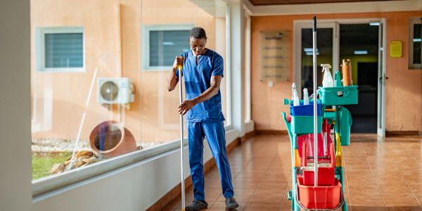 A janitor in blue scrubs mopping a hallway floor with a cleaning cart nearby.