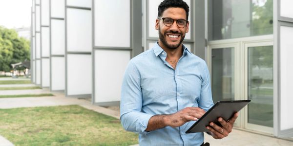 Smiling man in glasses using a tablet outdoors near modern buildings.