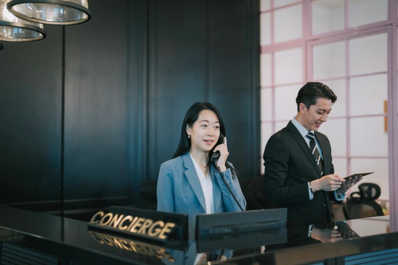 Asian hotel concierge team at the reception counter—female staff answering a phone call while her male colleague uses a digital tablet. Depicts professional hospitality service in a sleek, modern hotel setting. Ideal for themes related to customer service, digital check-in, and business travel.hotel receptionist, front desk, concierge service, hotel staff, Asian professionals, business attire, modern hotel, hospitality industry, customer service, digital check-in, smiling receptionist, teamwork, luxury hotel, business travel, front desk counter, lobby area, corporate hospitality, digital tablet, technology in hospitality, welcoming service,