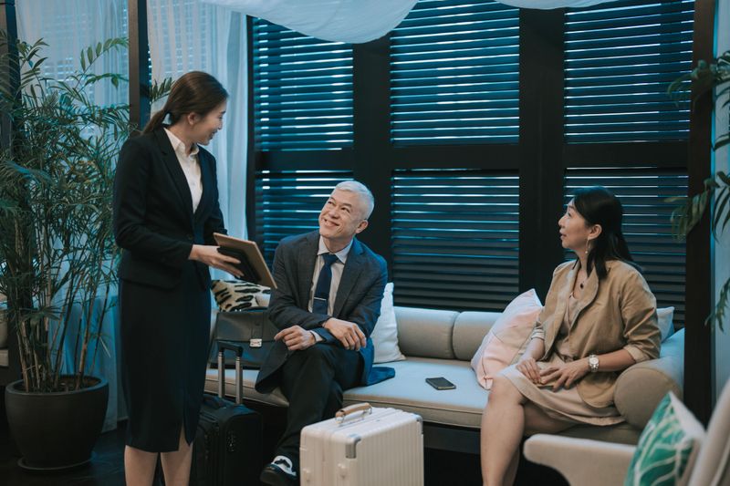 A professional Asian concierge in formal attire assists a businessman and a businesswoman during the hotel check-in process in a stylish hotel lounge. The concierge is using a digital tablet while guests, seated with their luggage, attentively review the information. The setting reflects modern hospitality, premium service, and business travel. Ideal for themes such as concierge service, hotel check-in, business travel, luxury hospitality, and customer experience.