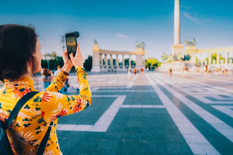 Tourist woman in a bright floral shirt stands in a spacious city square, capturing a photo of a historic monument with her smartphone. The scene is filled with sunlight and vibrant colours, reflecting the excitement of sightseeing and urban exploration on a beautiful day.