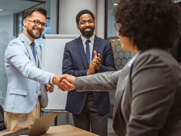 Two professionals shaking hands while a colleague applauds in a modern office.