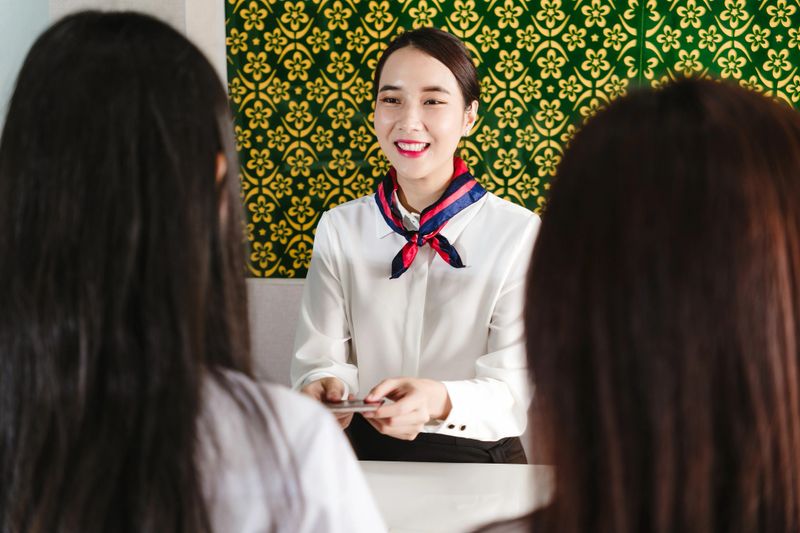Smiling airline ground staff hands documents to two female passengers at check-in desk. Depicts friendly customer service, travel preparation, and passenger assistance in modern airport.