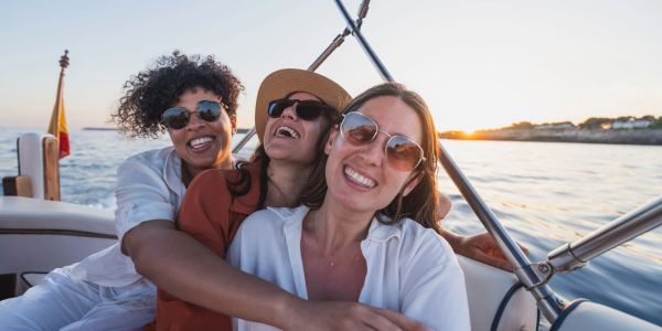 Three friends smiling and hugging on a boat at sunset.