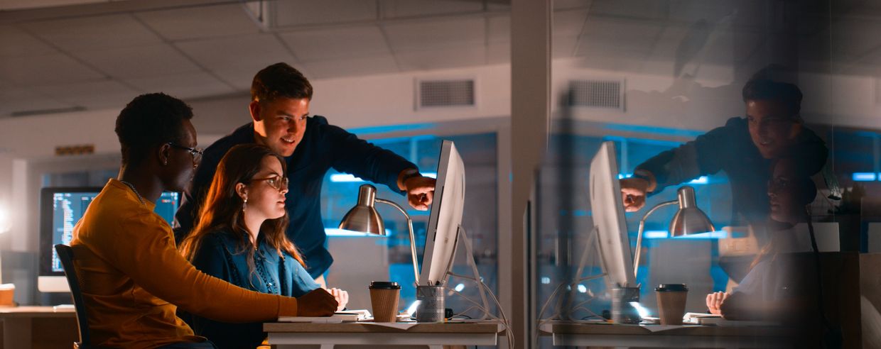 Three colleagues collaborate late at night, focused on a computer screen in an office.