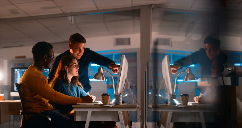 A group of diverse young professionals collaborate on a coding project in a modern office setting during nighttime. The vibrant atmosphere highlights teamwork, innovation, and dedication to achieving goals in a work environment.