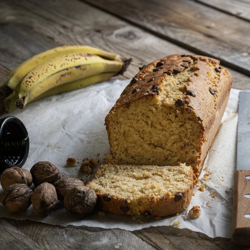 freshly made and sliced homemade banana bread on a rustic wooden table with fresh fruit and walnuts and kitchen utensils and natural elements. Natural light, pastries and artisanal breakfast