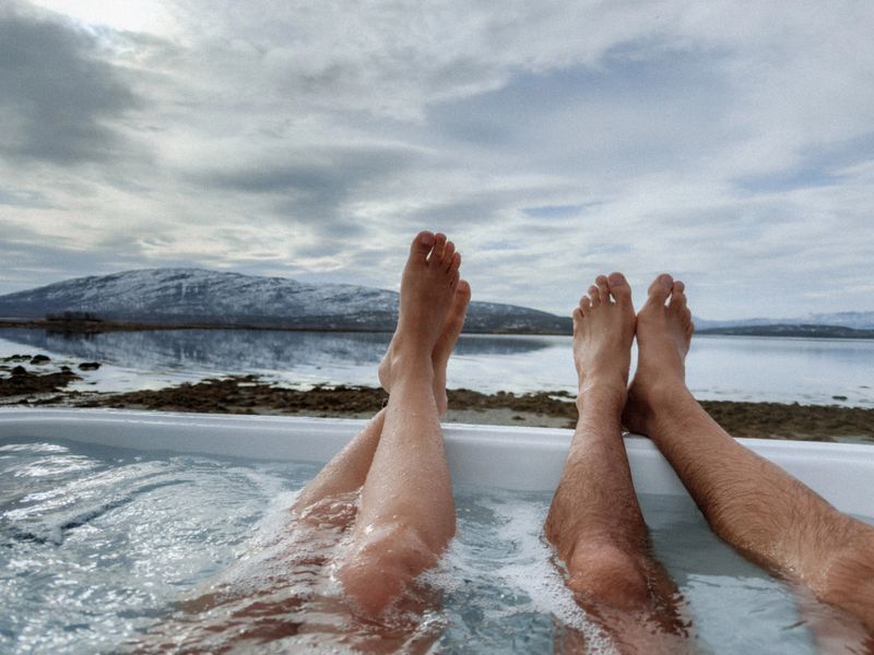 Couple's feet in hot tub overlooking tranquil mountain lake. Peaceful outdoor spa experience in nature. Scenic and serene relaxation setting.