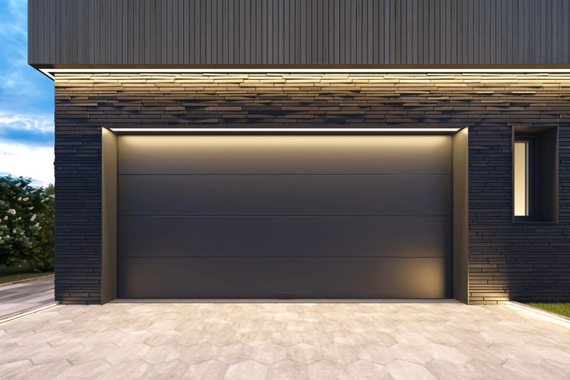 Front-facing view of a contemporary double garage, designed for two vehicles, featuring dark grey to black sectional roll-up doors. The facade is accented with sleek black slate stone, adding texture and contrast to the clean, modern lines of the home. The entrance design is minimalist and functional, blending seamlessly with the overall architectural style. Ideal image for use in showcasing modern garage solutions, facade design, urban housing concepts, or high-end real estate and construction materials. LED lights.