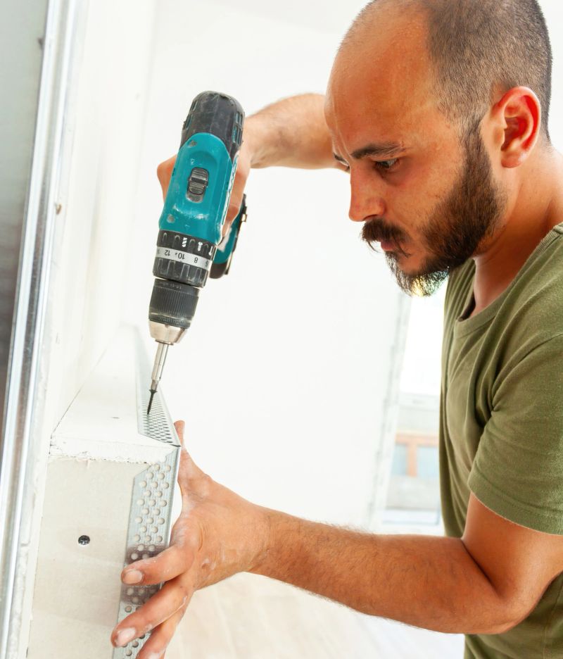 Construction worker is carefully installing a metal corner bead on a drywall using a cordless screwdriver, ensuring precise placement and secure fastening for a professional finish