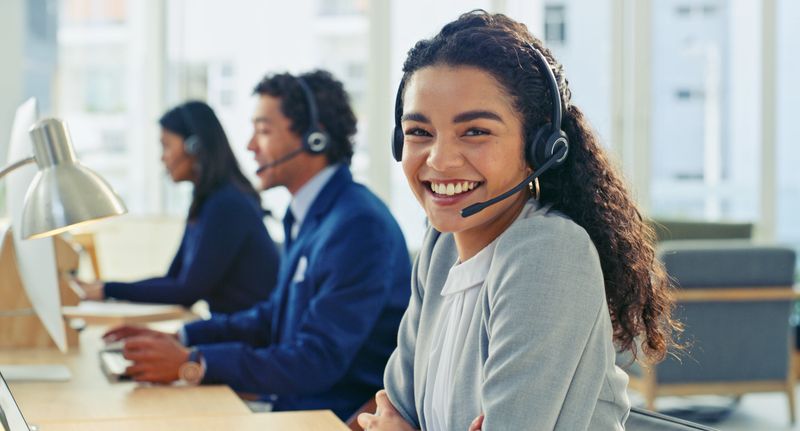 A cheerful customer support professional smiling at a desk with colleagues in a bright, open office. They wear headsets, working collaboratively to assist clients in a professional and welcoming atmosphere.