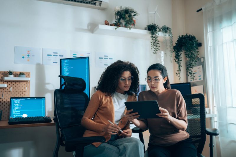 Women in tech collaborate on UI review with tablet and coding monitors in background