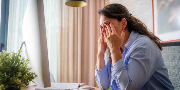 Woman in blue shirt rubbing her tired eyes at a desk with a computer.