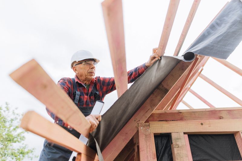 A professional builder carefully rolls out and secures waterproof roofing underlayment across wooden trusses during residential construction