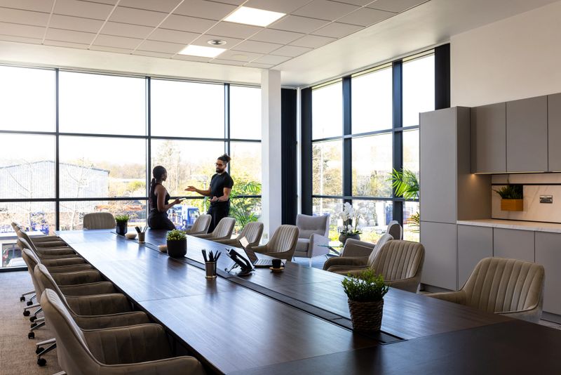 A bright and contemporary office conference room featuring a long meeting table surrounded by cushioned swivel chairs. Two business professionals engage in conversation near the large floor-to-ceiling windows that offer a view of an industrial complex and trees outside. The room is decorated with potted plants and minimalist cabinetry, creating a clean and professional atmosphere ideal for corporate meetings or collaborative work.

Videos are available similar to the scenario