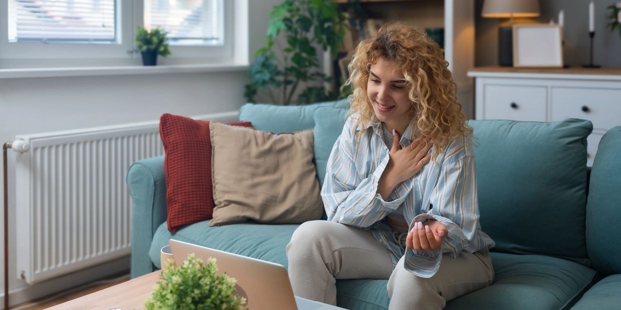 A woman smiling and touching her chest during a video call at home.