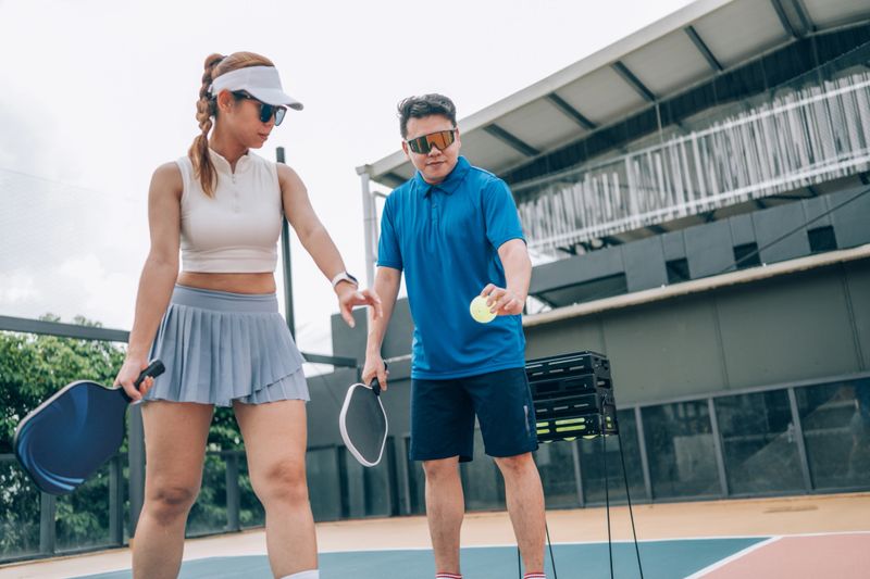 An Asian male pickleball coach is teaching a Asian woman to play pickleball on an outdoor court.