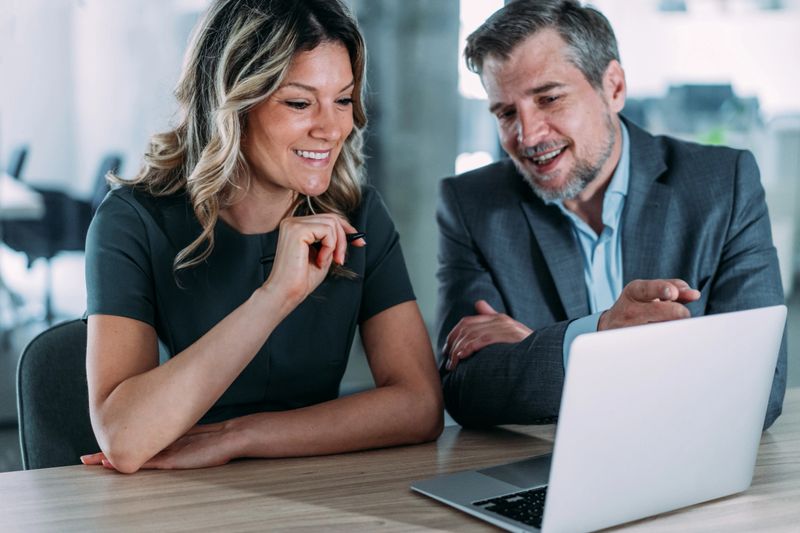 Shot of a two confident business persons sitting on a desk in the office and sharing ideas. Businessman and businesswoman in meeting using laptop and discussing business strategy. Business coworkers working together in the office.