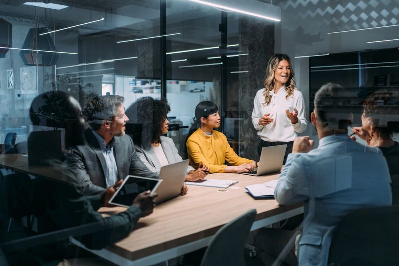 Shot of a businesswoman delivering a speech during a meeting in conference room. Businesswoman leading a training class for professionals. Businesswoman talking on seminar in front of her colleagues. Group of business persons in business meeting. Group of entrepreneurs on meeting in board room. Creative business team on meeting in the office. The view is through glass.