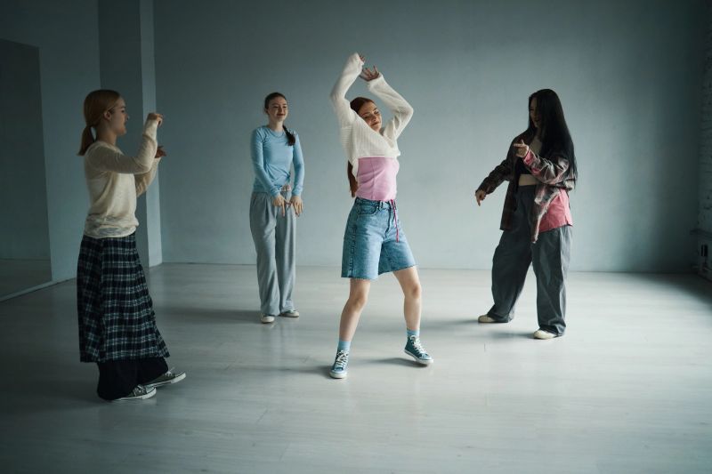 Group of young people practicing dance moves in spacious studio with gray walls, wearing casual clothes. People smiling and having fun while engaging in activity