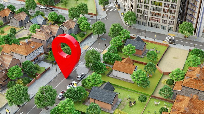 Digitally rendered illustration of a suburban neighborhood with houses, trees, and streets. The central focus is a large, red map location pin marker hovering over a street, symbolizing a geolocation or navigation point - commonly associated with GPS, maps, or location-based services. The setting is clean and modern, with a mix of single-family homes and urban buildings in the background, emphasizing the integration of digital tools with real-world environments.