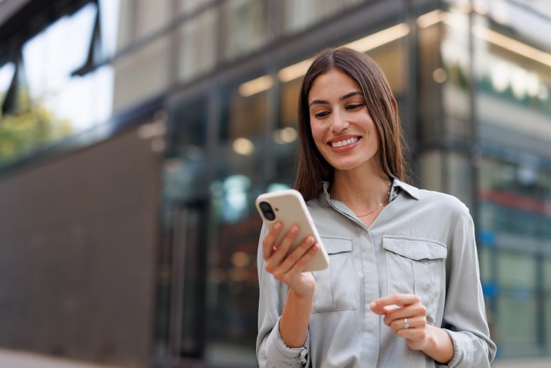 Young businesswoman smiling while using her smartphone, enjoying connectivity in front of sleek modern office buildings in the urban landscape