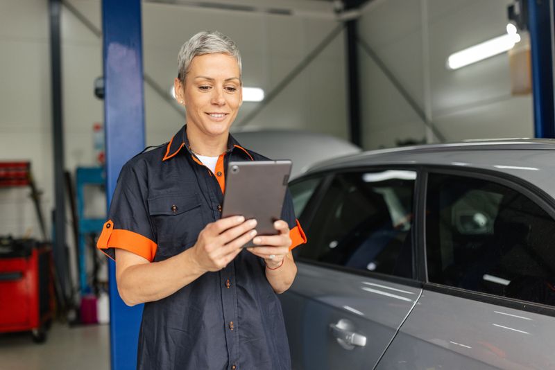 A professional female mechanic stands with a tablet beside a parked car in an organized garage, indicating readiness and expertise in providing automotive solutions.