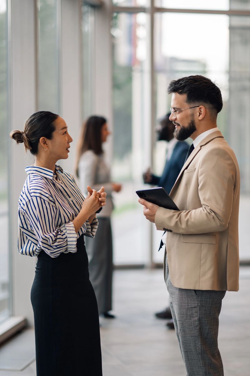 Businessman and businesswoman discussing work while holding a tablet in a bright, modern office lobby with large glass windows