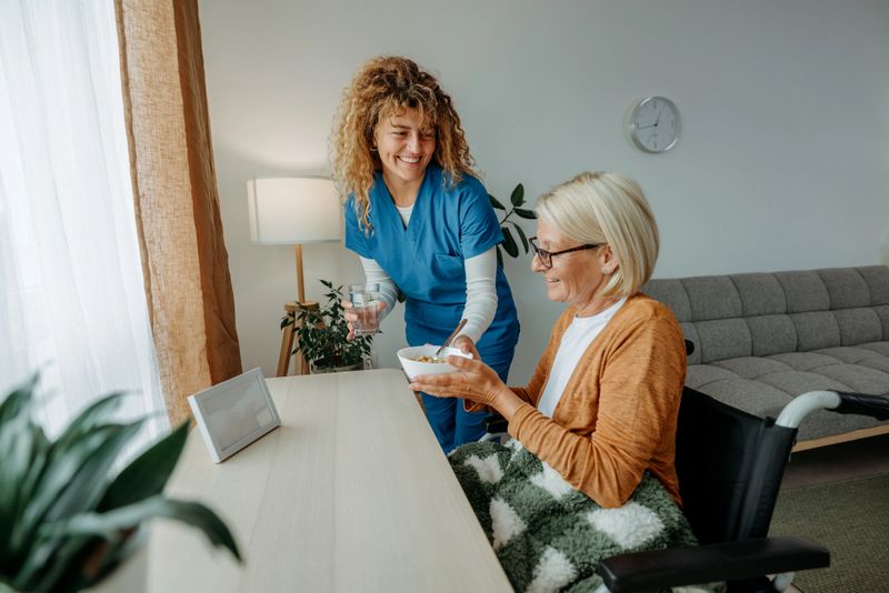 Caring female healthcare worker delivering breakfast tray to a senior woman sitting in a wheelchair. Supportive and attentive home care in a warm, domestic environment.