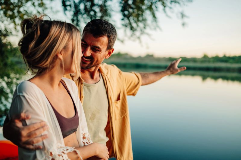 Young adult couple embracing by a tranquil lake at sunset, enjoying the serene natural landscape. The man gestures towards the horizon, capturing a moment of connection