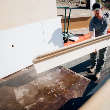 Man carrying a long wooden plank outdoors near a building.