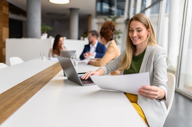 Female lawyer analyzing documents and using laptop while working in conference room with colleagues in the background