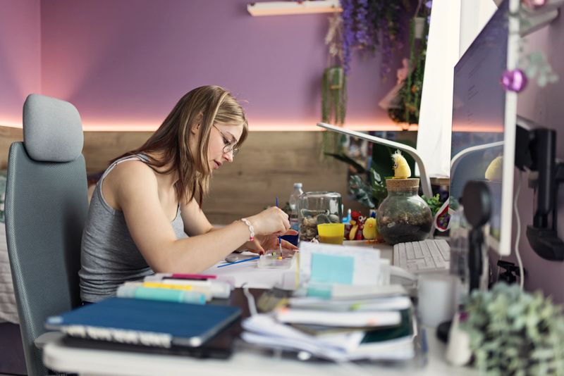 A young woman is seated at a desk in a cozy room, deeply focused on her arts and crafts project. She is holding a paintbrush and appears to be mixing paint for painting. The desk is cluttered with various art supplies, including paint tubes and brushes. A jar filled with seashells and other decorative items is also visible on the desk. The room has a warm ambiance with soft lighting and wooden walls, creating a comfortable and inviting atmosphere for creative work.
Shot with Canon R5