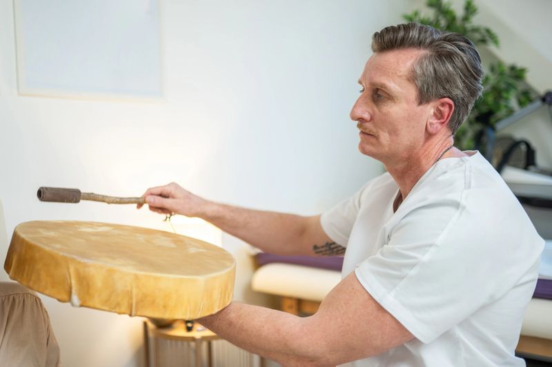 A mid-aged Caucasian male therapist wearing a white shirt uses a shamanic drum in an indoor therapeutic space surrounded by soft lighting and natural decor.
