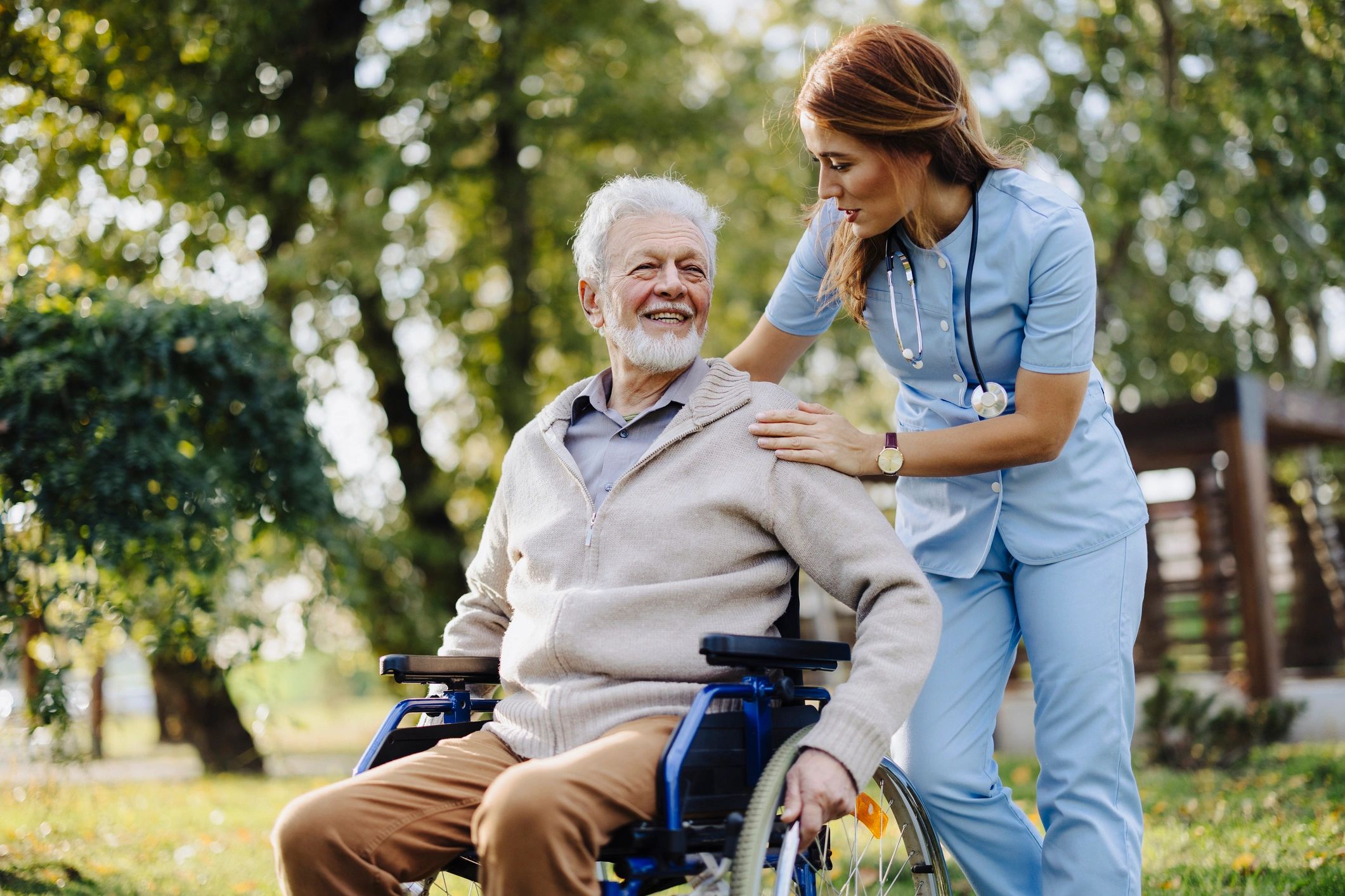 A female caregiver in blue scrubs talking to an elderly man in a wheelchair.