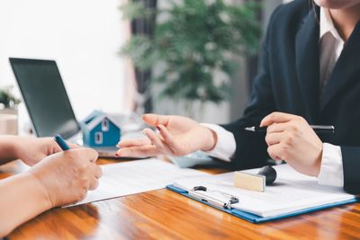 Two people discussing and signing a contract at a wooden desk with a laptop and a house model.