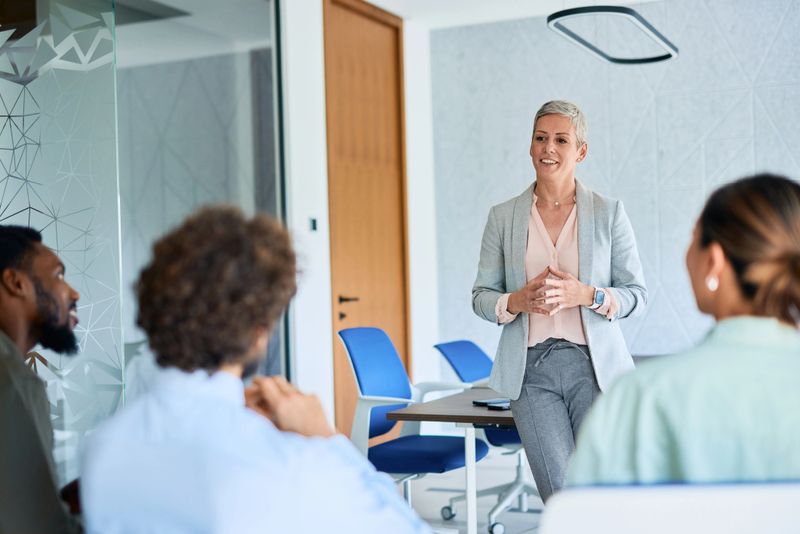 Mature businesswoman is standing and leading a meeting with her team, discussing new business ideas and strategies in a modern office environment