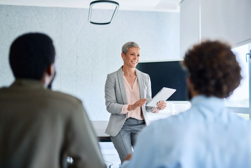 Mature businesswoman leads a professional meeting, engaging with her team while holding a digital tablet and presenting information in a modern office setting