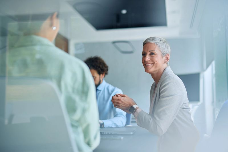 Businesswoman is smiling while listening to a colleague during a business meeting in a modern office, her colleagues are sitting at the table and listening