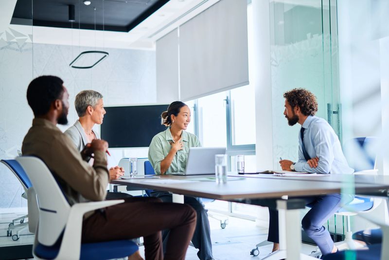 Four businesspeople sitting around a table in a modern office, engaging in a project discussion, with a woman gesturing animatedly while sharing her ideas