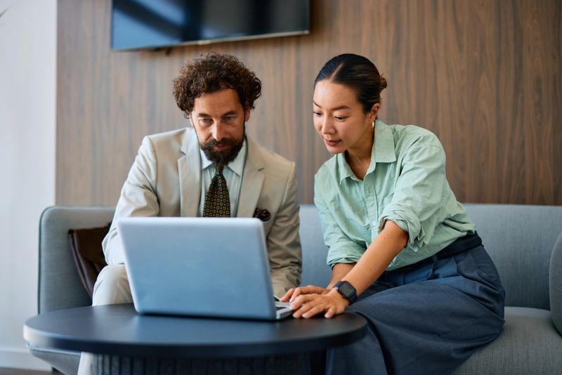 Two business partners sitting on a modern office sofa, collaborating on a laptop, discussing project details, and sharing ideas for successful strategies and innovative solutions