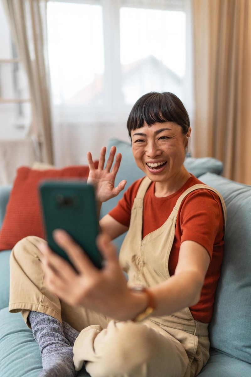 Smiling mature asian woman waving while enjoying a video call on her smartphone, comfortably seated on the sofa in her cozy living room, radiating happiness and connection
