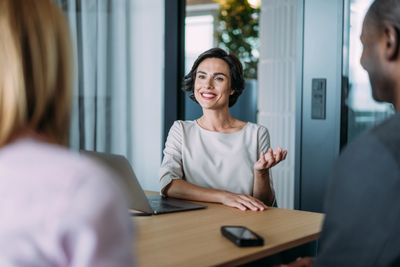 A woman in a white blouse is smiling and talking during a meeting with two colleagues.