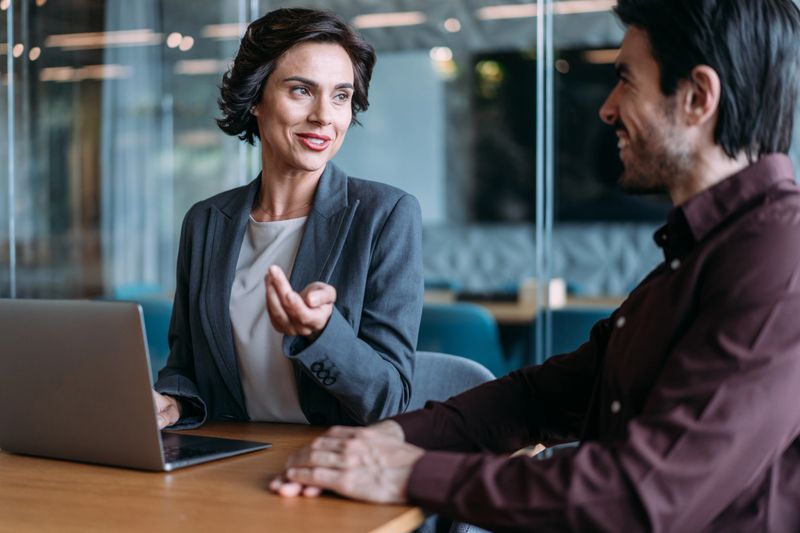 Shot of a two confident business persons sitting on a desk in the office and sharing ideas. Businessman and businesswoman in meeting using laptop and discussing business strategy. Business coworkers working together in the office.
