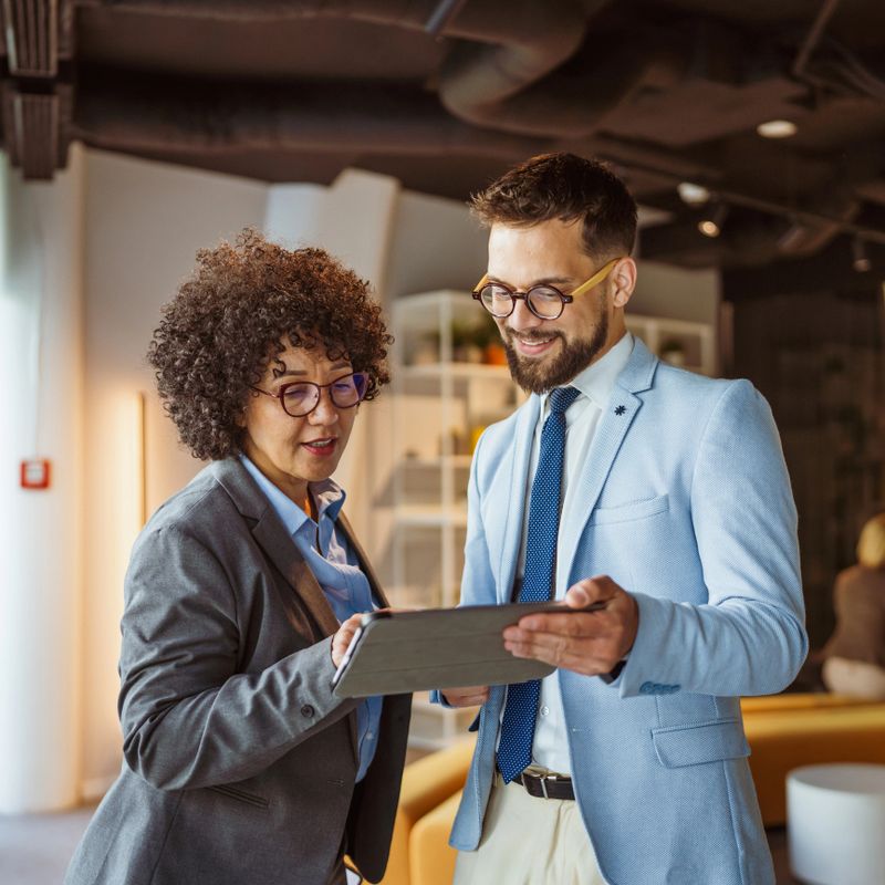 Couple of mature adult male and female colleagues walk in the office use tablet and discuss work. Motivated diverse business people in modern open office