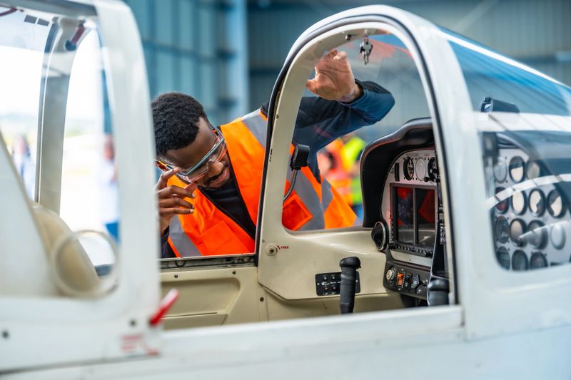 Aircraft engineer wearing safety glasses and a high-visibility vest inspects the cockpit of a small plane inside an aircraft hangar