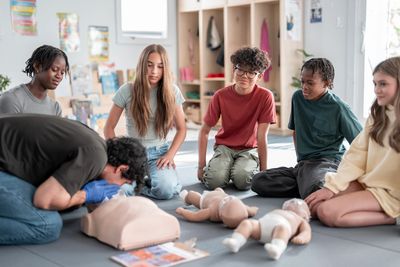 Kids attentively watch CPR training using adult and infant mannequins.