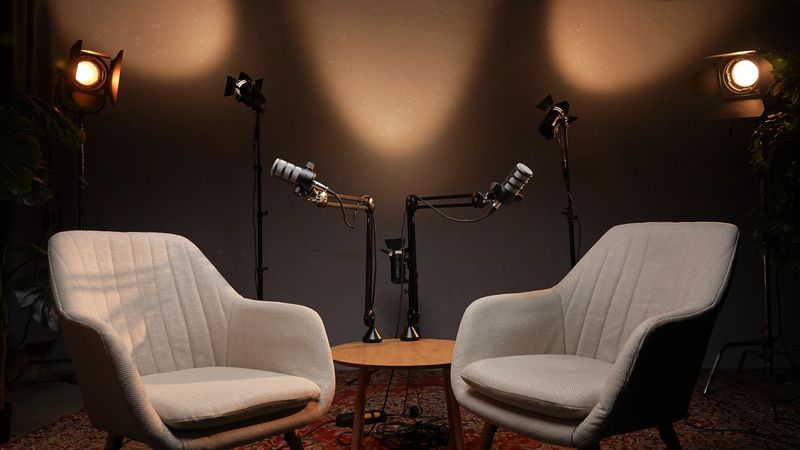 In a thoughtfully designed podcast studio, two stylish chairs face each other across a table, with microphones poised for an engaging interview. Soft light highlights the inviting space.