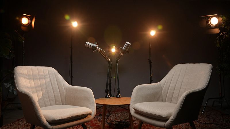 Two comfortable chairs are positioned facing each other in a cozy podcast studio, illuminated by warm lights. This inviting setup is perfect for insightful interviews on various topics.